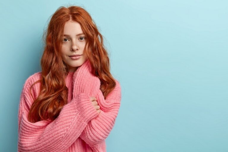 Waist up shot of lovely teenager with long red hair, has direct gaze at camera with serious expression, no make up, dressed in oversized knitted jumper, isolated over blue background, empty space