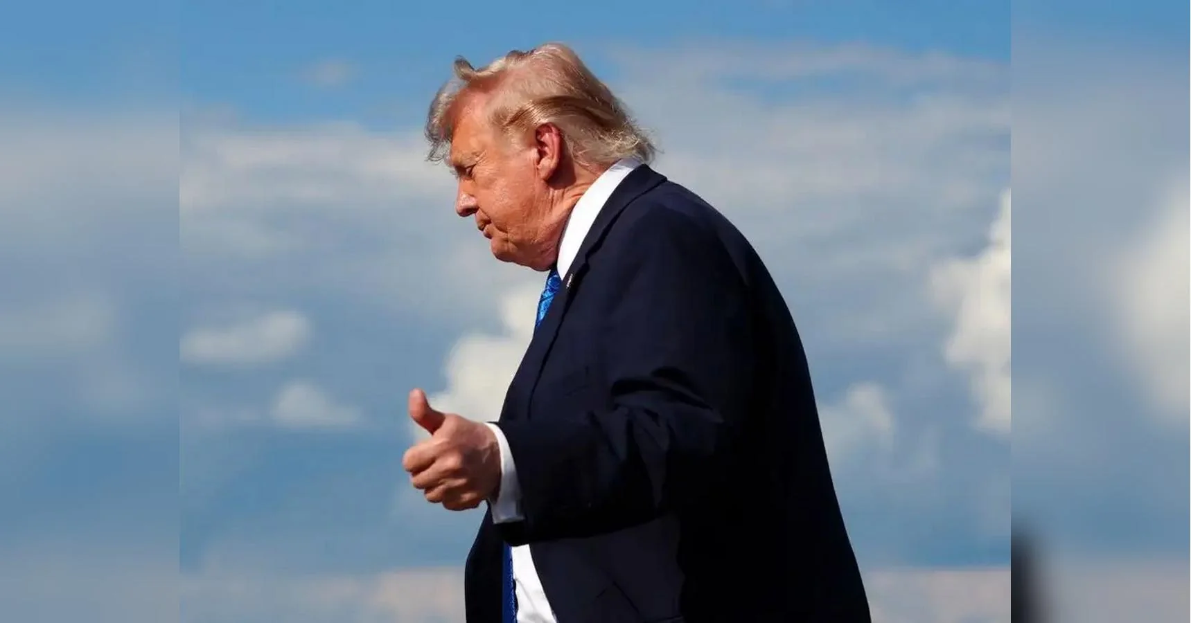 Дональд Трамп у темному костюмі показує великий палець вгору на тлі блакитного неба