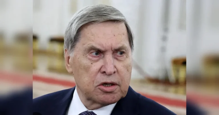 Літній чоловік у темному костюмі говорить у парадній залі з червоною доріжкою