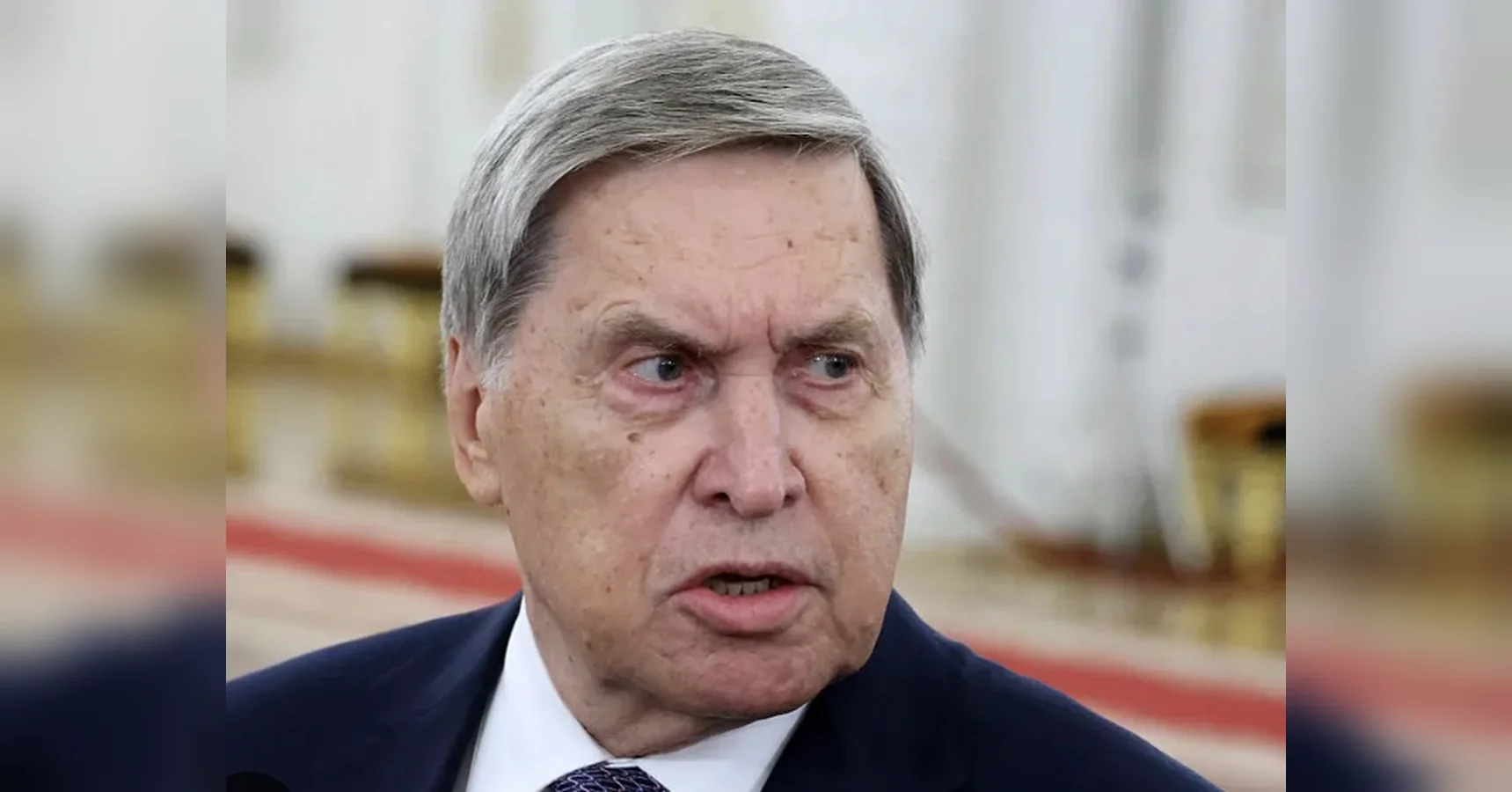 Літній чоловік у темному костюмі говорить у парадній залі з червоною доріжкою