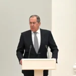 Сергій Лавров у костюмі виступає за білою трибуною з мікрофонами