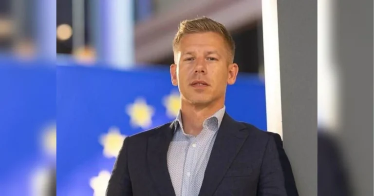 Чоловік у темному піджаку стоїть біля стіни на тлі прапора ЄС