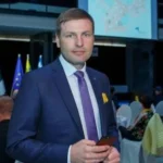 Чоловік у синьому костюмі з фіолетовою краваткою тримає телефон на офіційному заході з прапорами