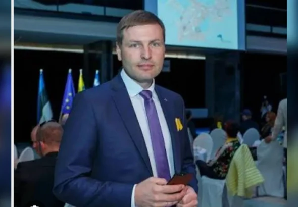 Чоловік у синьому костюмі з фіолетовою краваткою тримає телефон на офіційному заході з прапорами