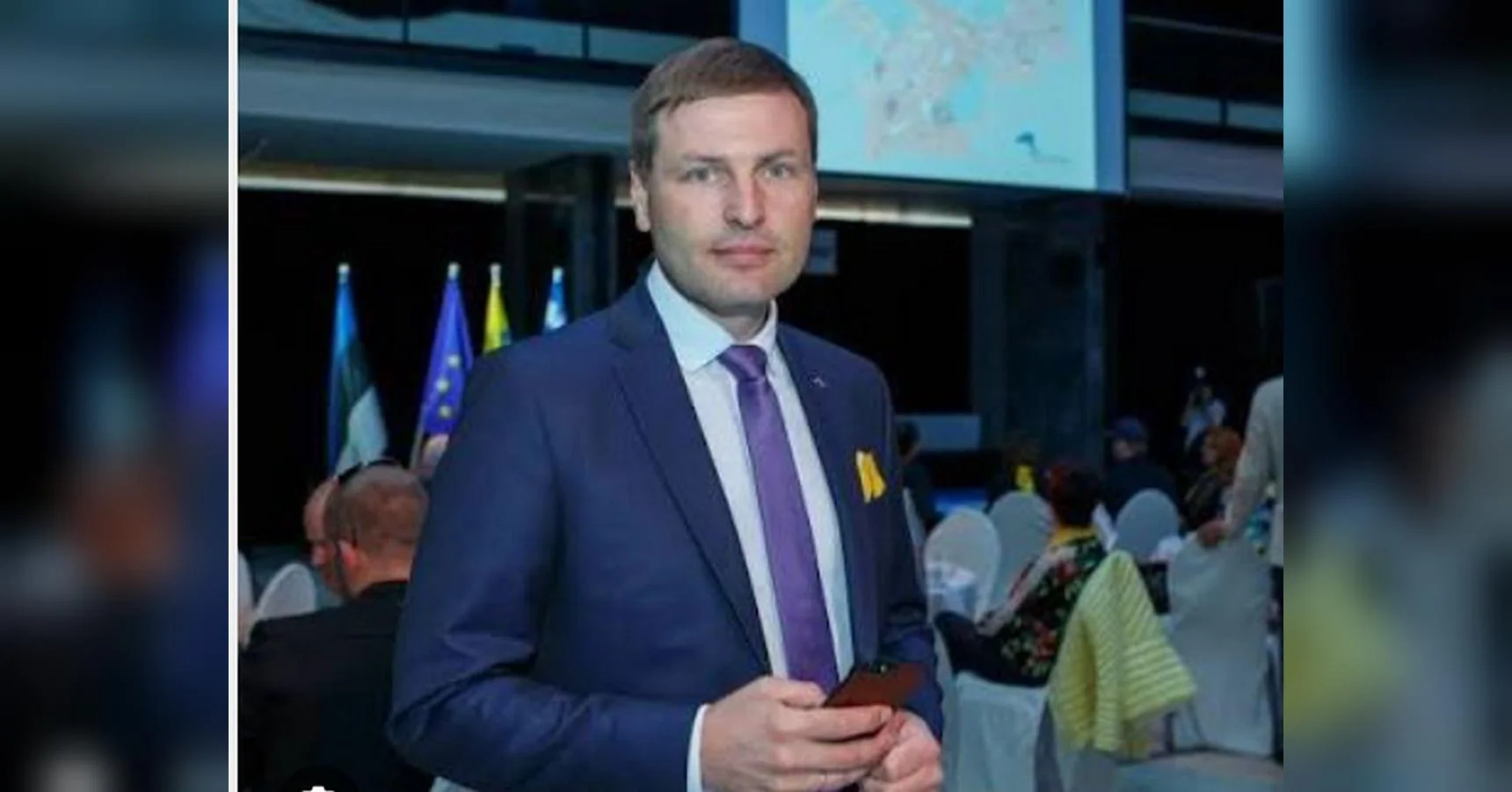 Чоловік у синьому костюмі з фіолетовою краваткою тримає телефон на офіційному заході з прапорами