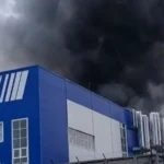 Синьо-білий заводський корпус з трубами, над яким клубочиться густий чорний дим