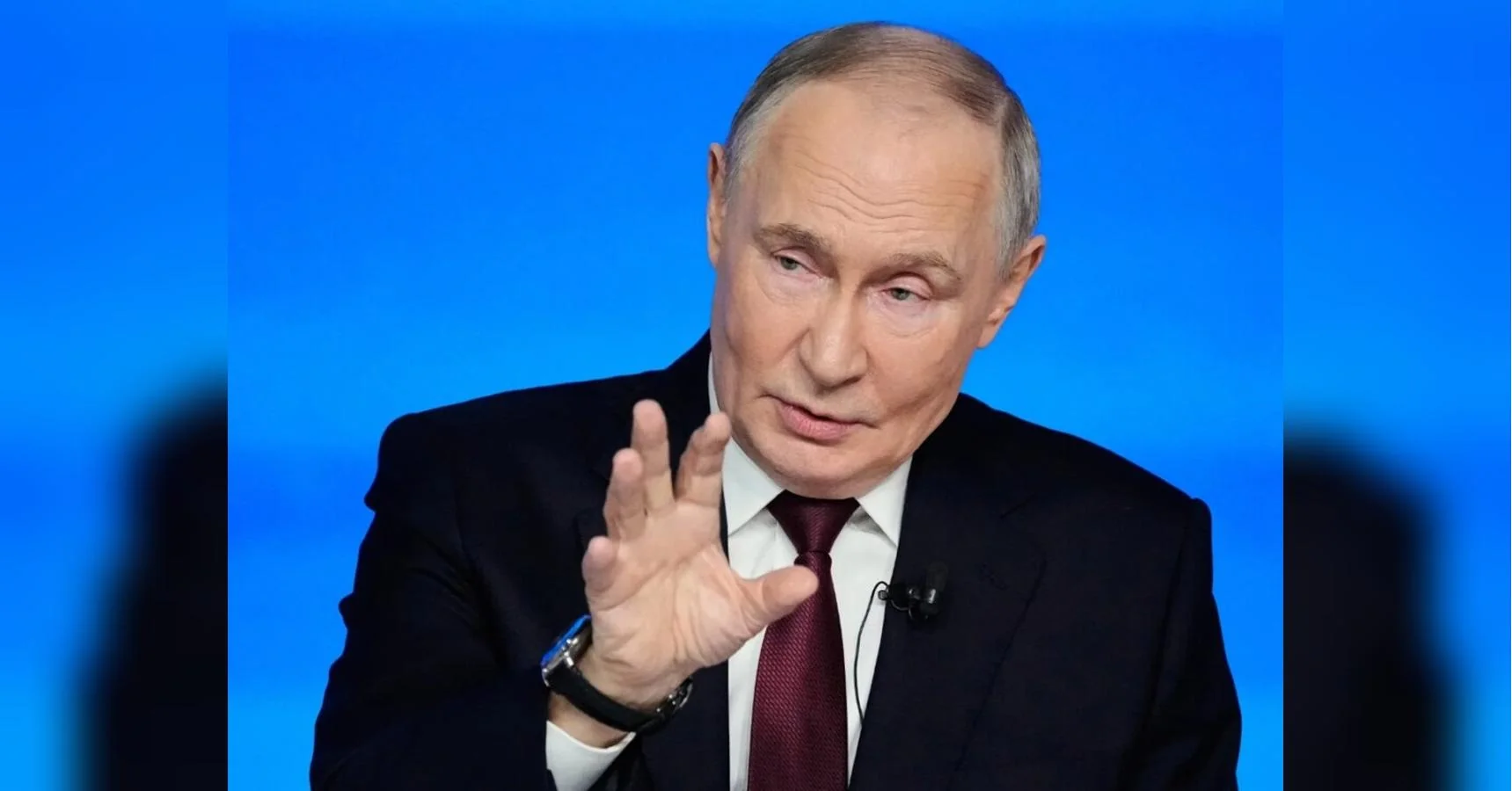 Володимир Путін у костюмі жестикулює рукою на синьому тлі