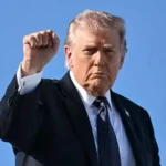 Дональд Трамп у костюмі піднімає кулак на тлі блакитного неба