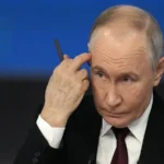 Володимир Путін у костюмі тримає ручку біля скроні на синьому тлі